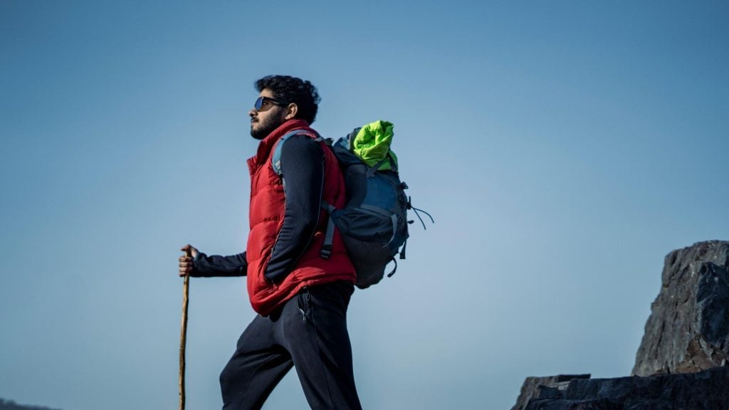 A man wearing a blue shirt, red vest, backpack, and sunglasses hiking with a walking stick