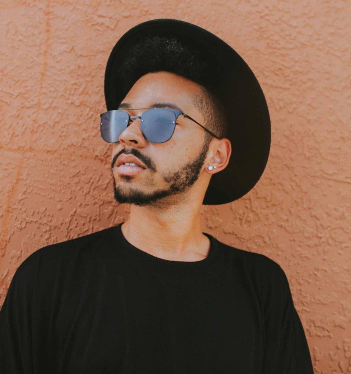 A man wearing a black T-shirt, wide-brimmed hat, and oversized black sunglasses