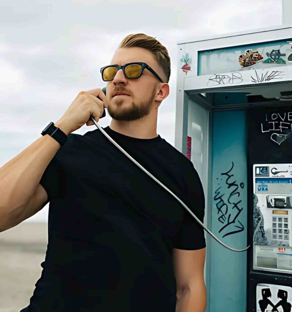 A man in a black T-shirt and black-framed yellow-tinted sunglasses using an old graffiti-covered payphone