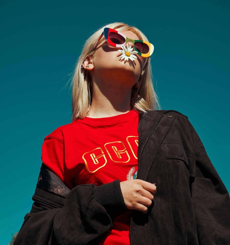 Tan blonde woman in multicolor striped cat-eye sunglasses, a red shirt, and black suede jacket