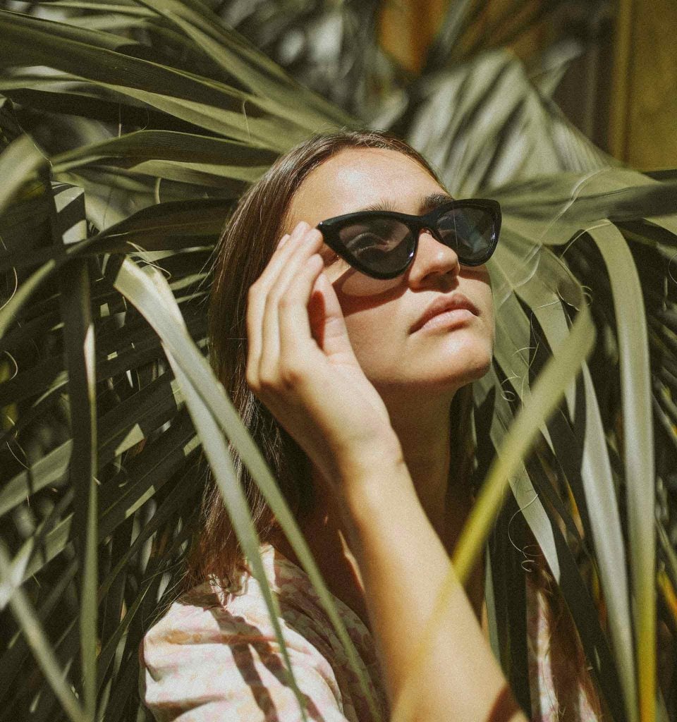 Woman in black cat-eye sunglasses surrounded by palms as she stares up at the sky