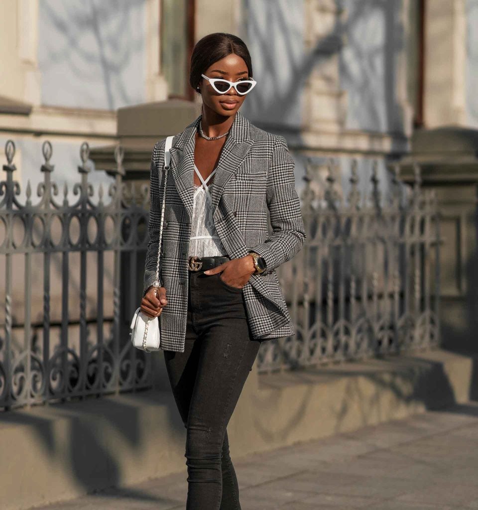 Chic woman strolling down a sidewalk wearing black jeans, white bustier, herringbone blazer, and white cat-eye sunglasses