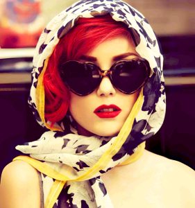 A woman with striking red hair wearing tortoiseshell heart sunglasses and a vintage floral scarf, creating a fabulous retro look