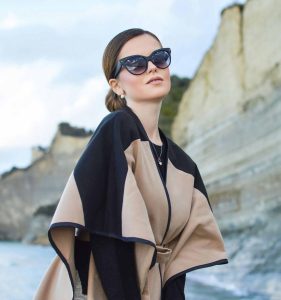 A woman wearing stylish cape and oversized cat-eye sunglasses while standing in front of the sea