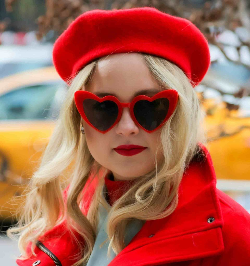 A fashionable blonde woman clad in a red wool jacket, red beret, and oversized red-framed heart sunglasses