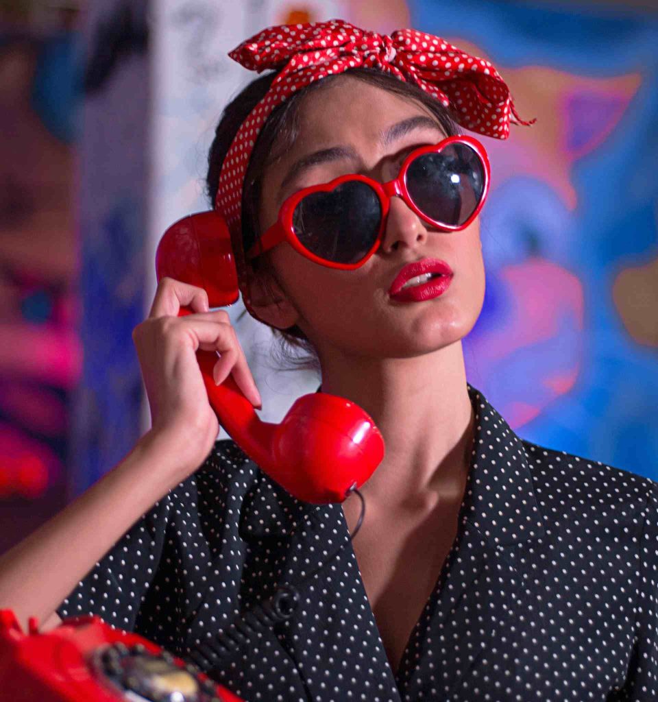 A woman wearing a navy polka-dot dress, red polka-dot headscarf, and red heart sunglasses while talking on a red rotary phone