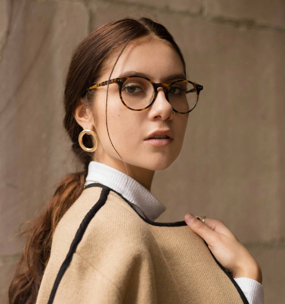 A young woman wearing tortoiseshell round glasses, gold circular earrings, a white turtleneck, and a beige and black Inverness cape