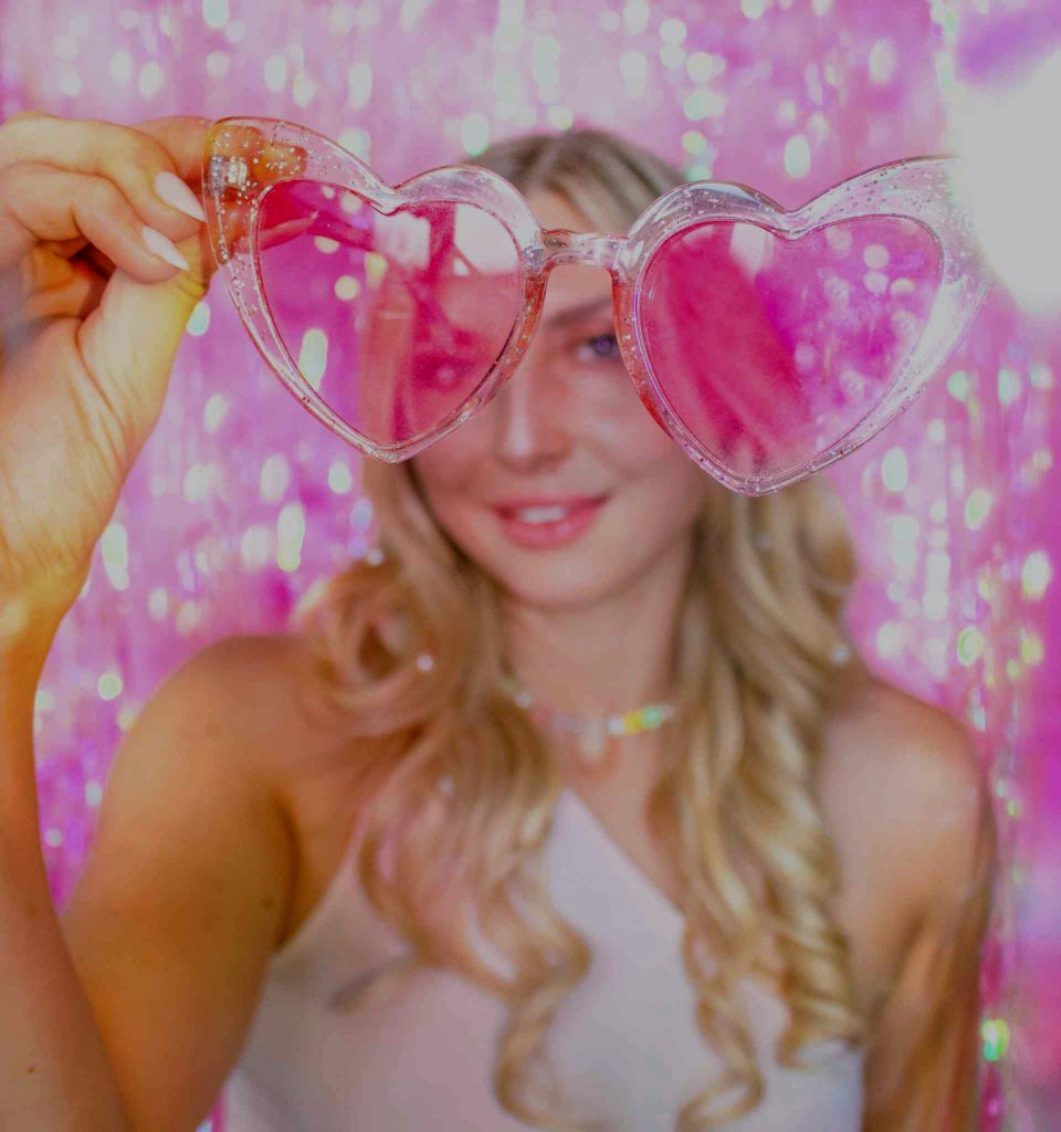 A blonde woman wearing a white off-the-shoulder dress and holding glittery heart-shaped sunglasses with pink lenses