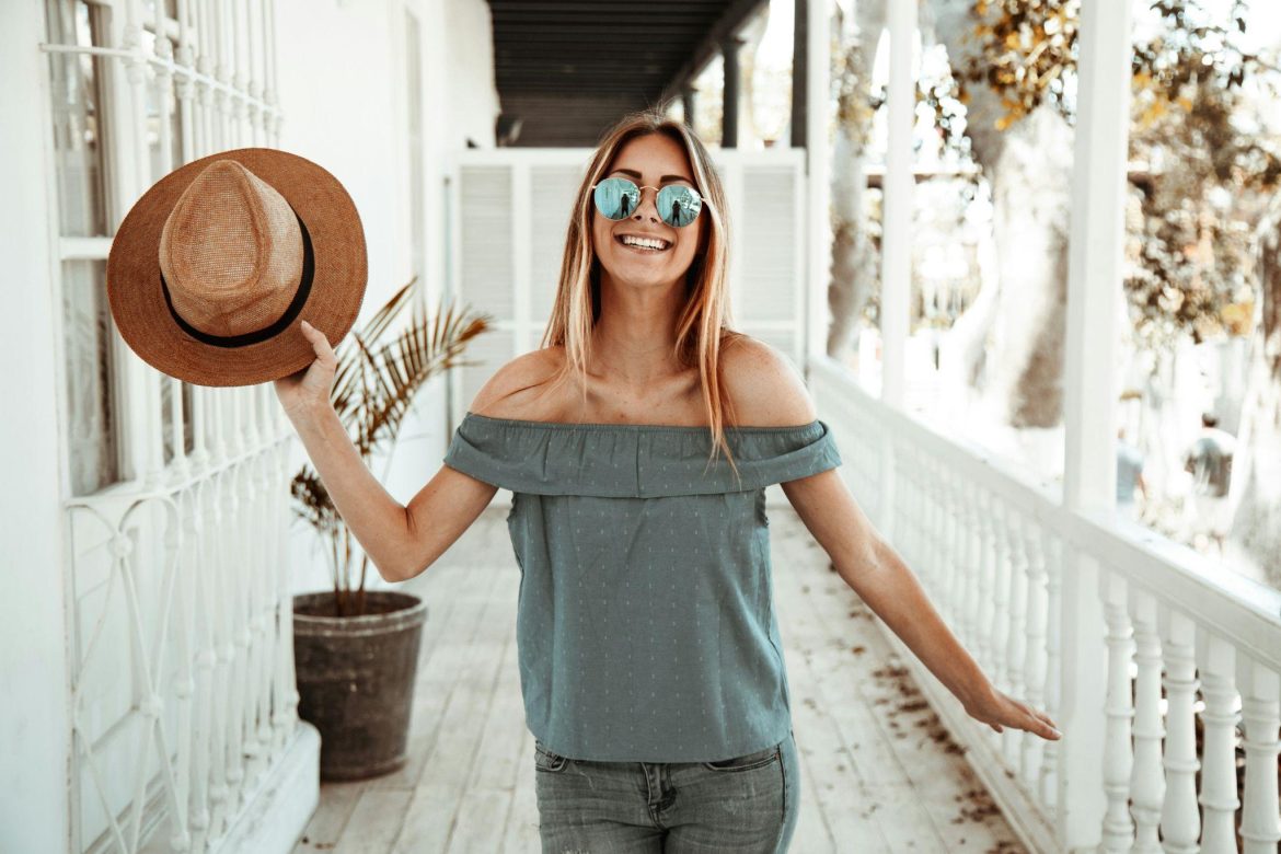 Woman smiling while wearing a grey off-the-shoulder blouse and blue tinted sunglasses