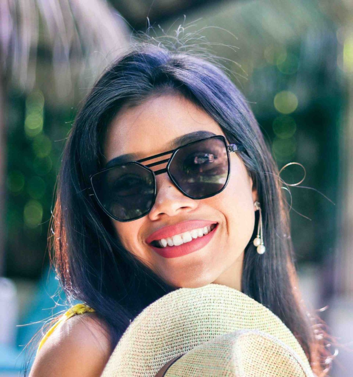 A woman in triple-bridged black sunglasses holding a straw hat and showing off her big smile on a sunny day