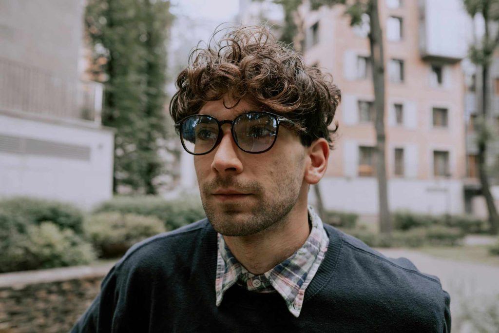 A man wearing a plaid button-down shirt, dark pullover, and glasses with dark, thin frames and clear lenses